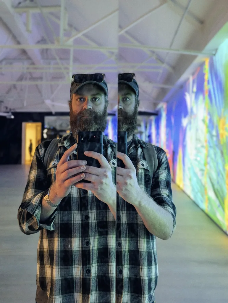 Featured image for Words about 2025 - Jay Springett taking a mirror selfie in a plaid shirt with a colorful light installation in the background.